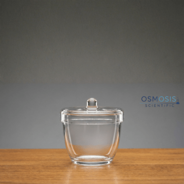 A transparent quartz laboratory crucible with a matching lid featuring a top knob, resting on a wooden surface.