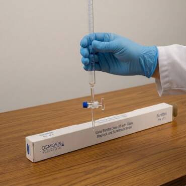 A laboratory technician in blue gloves holding a clear borosilicate glass burette with a blue Schellbach stripe and a glass stopcock, positioned over its Osmosis Scientific packaging.